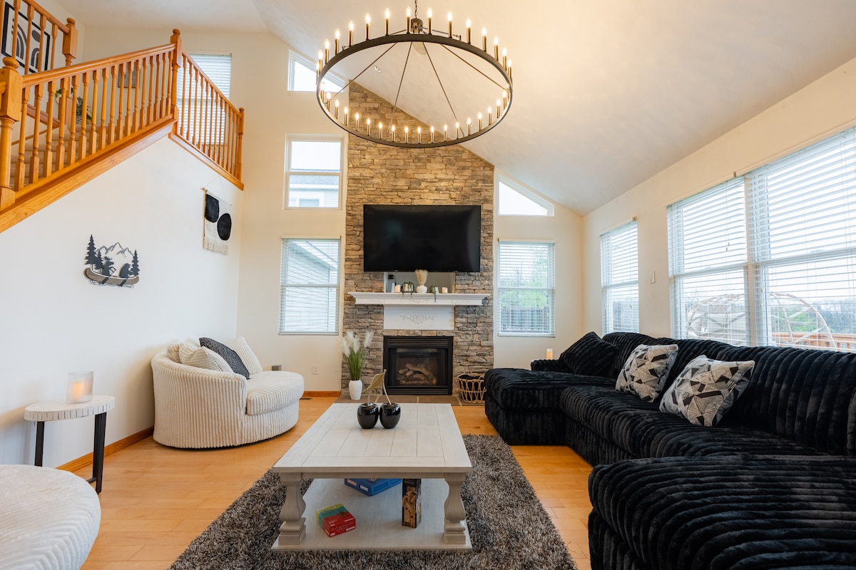 The grand living room features high vaulted ceilings, a stone fireplace, and a large 75-inch television. A spacious sectional couch sits invitingly, complemented by a cozy area rug and a stylish coffee table. Natural light floods through multiple windows, enhancing the open and airy atmosphere.