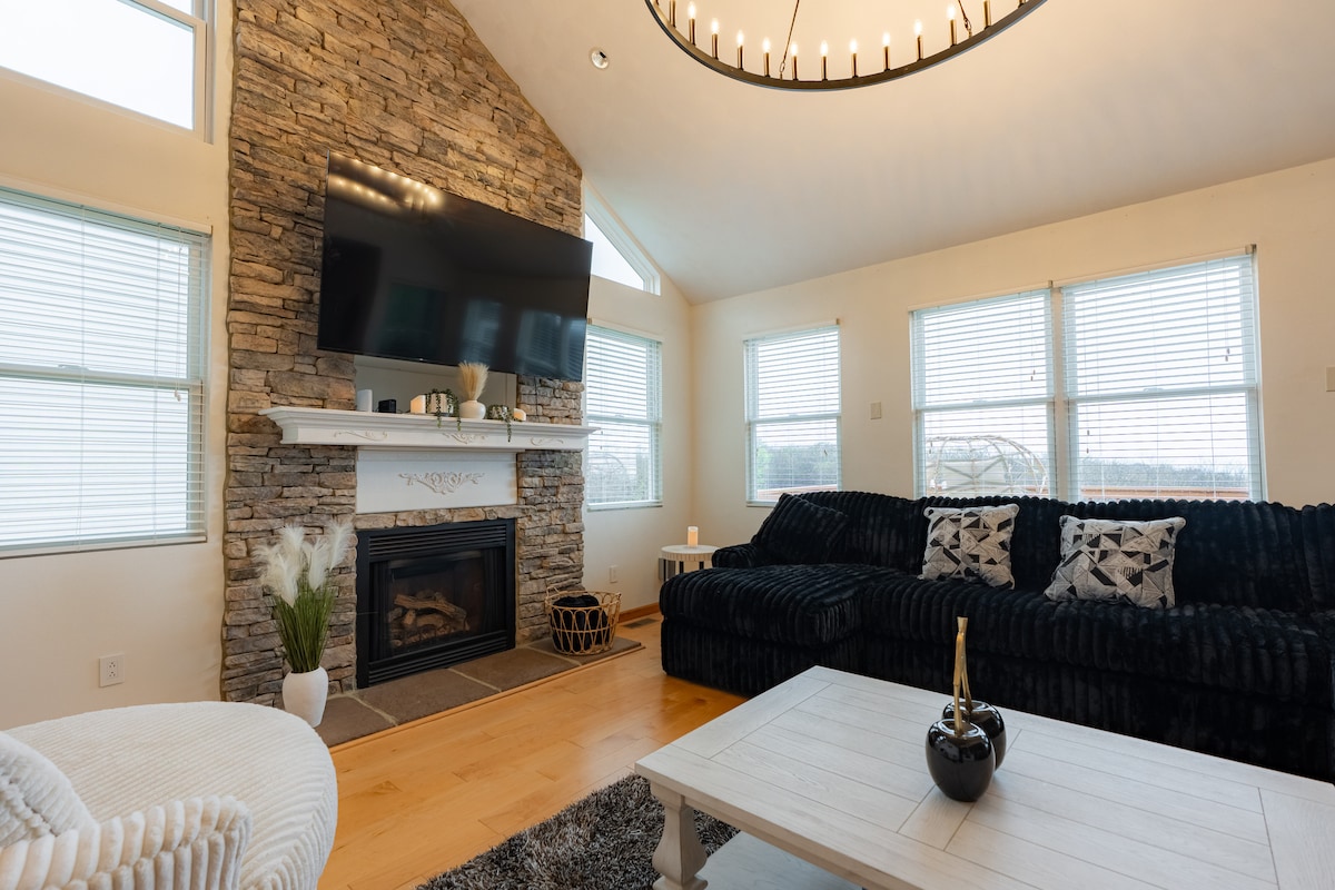 A spacious living room is highlighted by stone accents and vaulted ceilings. A large flat-screen TV is mounted above a modern fireplace. The comfortable seating area features dark plush sofas, and sunlight streams in through multiple windows, creating a bright and inviting ambiance.