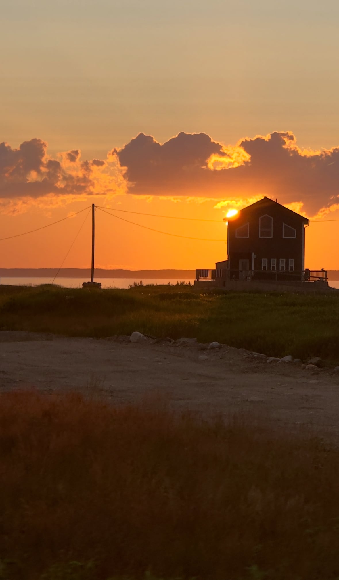 A silhouette of a charming house is captured against a vibrant sunset, with the sun partially hidden behind the structure. Soft clouds are visible in the sky, reflecting warm hues of orange and yellow. A grassy area and a dirt path are positioned in the foreground.