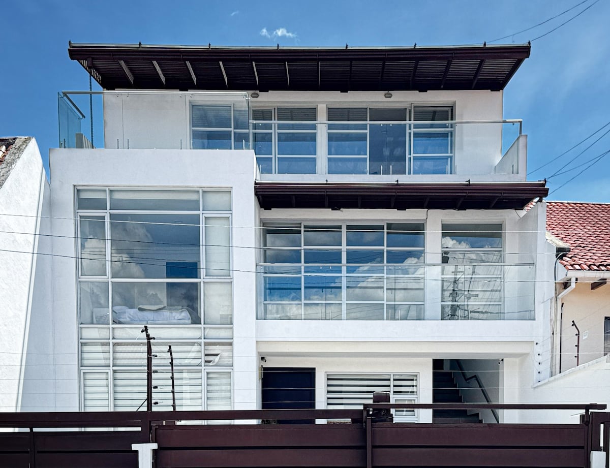The exterior of the building showcases a modern design with multiple large windows and balconies. A clean white facade is complemented by transparent railings, and the structure is set against a clear blue sky, enhancing its contemporary appearance.