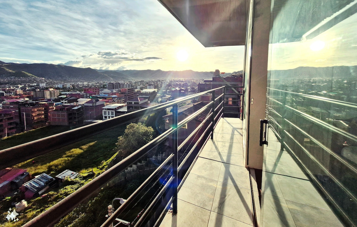A private balcony is visible, offering expansive city and mountain views. Glass panels reflect sunlight, creating a bright and open atmosphere. The balcony is equipped with a metal railing and tiled flooring, enhancing the sense of space and serenity.