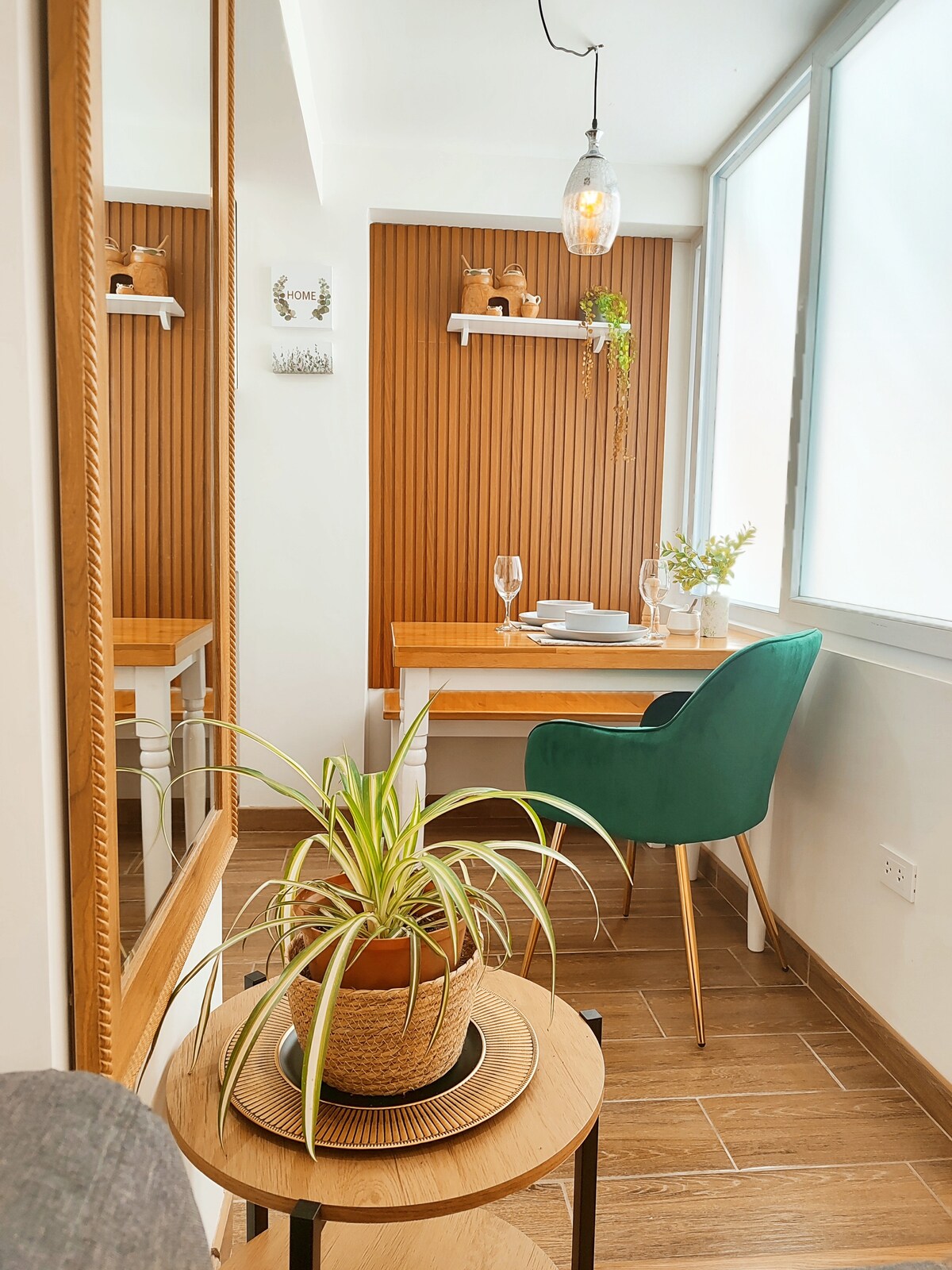 A dining area is featured with a wooden table and a single green chair. A potted plant decorates the table, while a mirror reflects the space. Warm wood paneling accentuates the walls, and a pendant light hangs overhead, providing soft illumination.