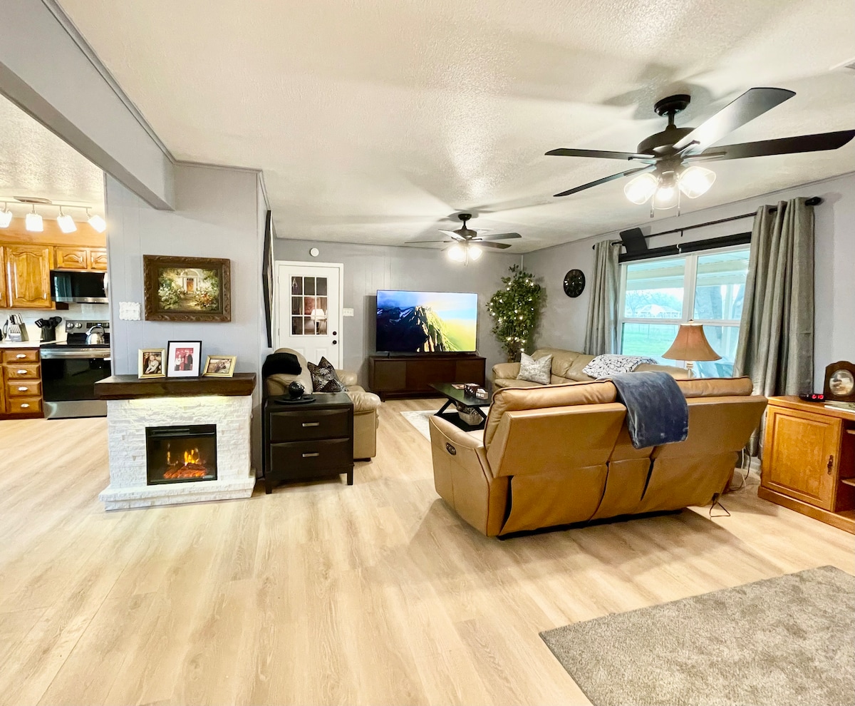 A comfortable living area is equipped with leather sofas and recliners, accommodating several guests. A large television is positioned against the wall, while a decorative fireplace adds warmth to the space. Natural light filters through a large window, enhancing the inviting ambiance.