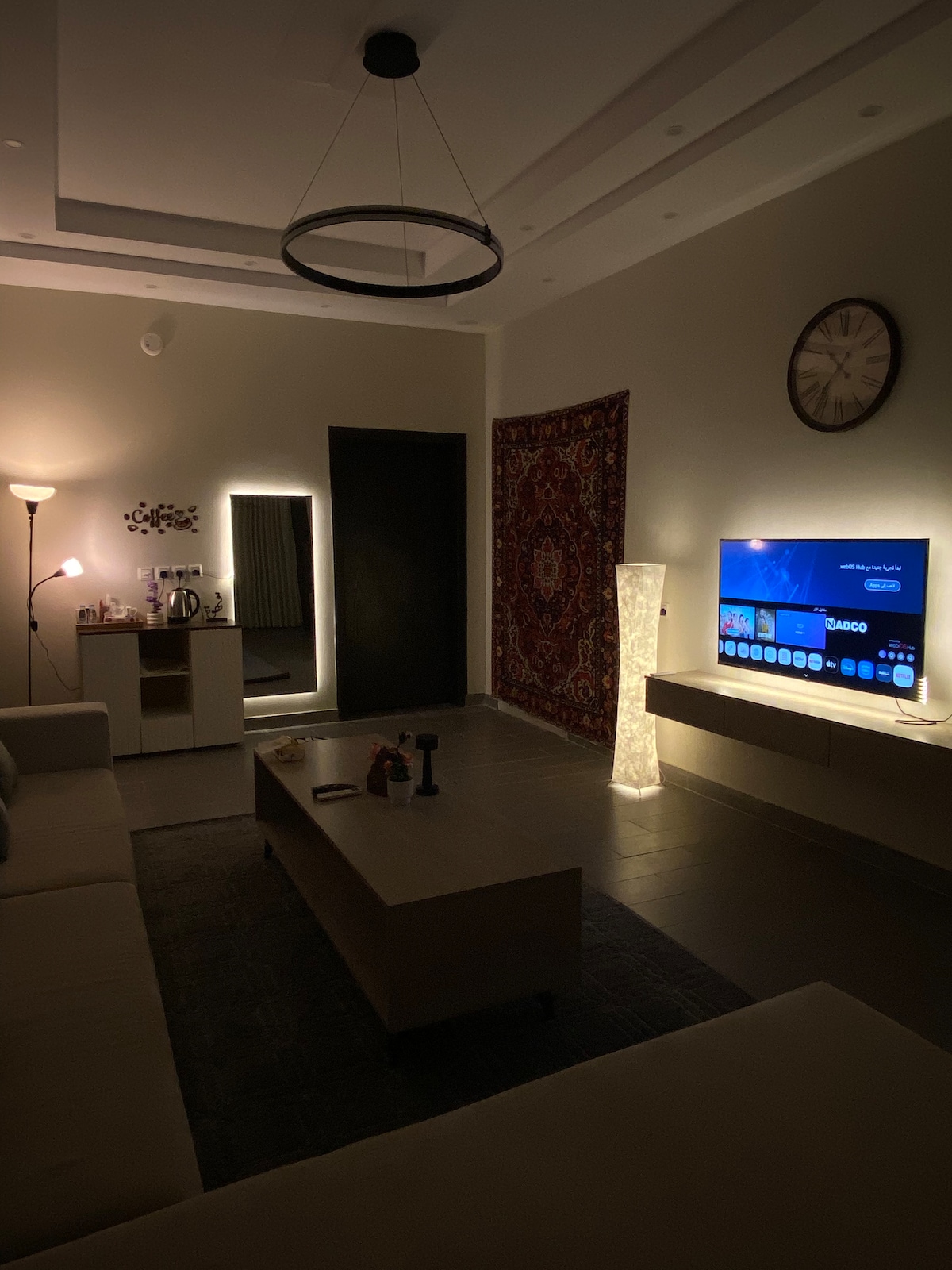 The living area features a neutral color palette with a spacious sofa facing a large television. A floor lamp and decorative light provide soft illumination. A mirror and an ornate rug add visual interest, while a clock on the wall indicates the time.