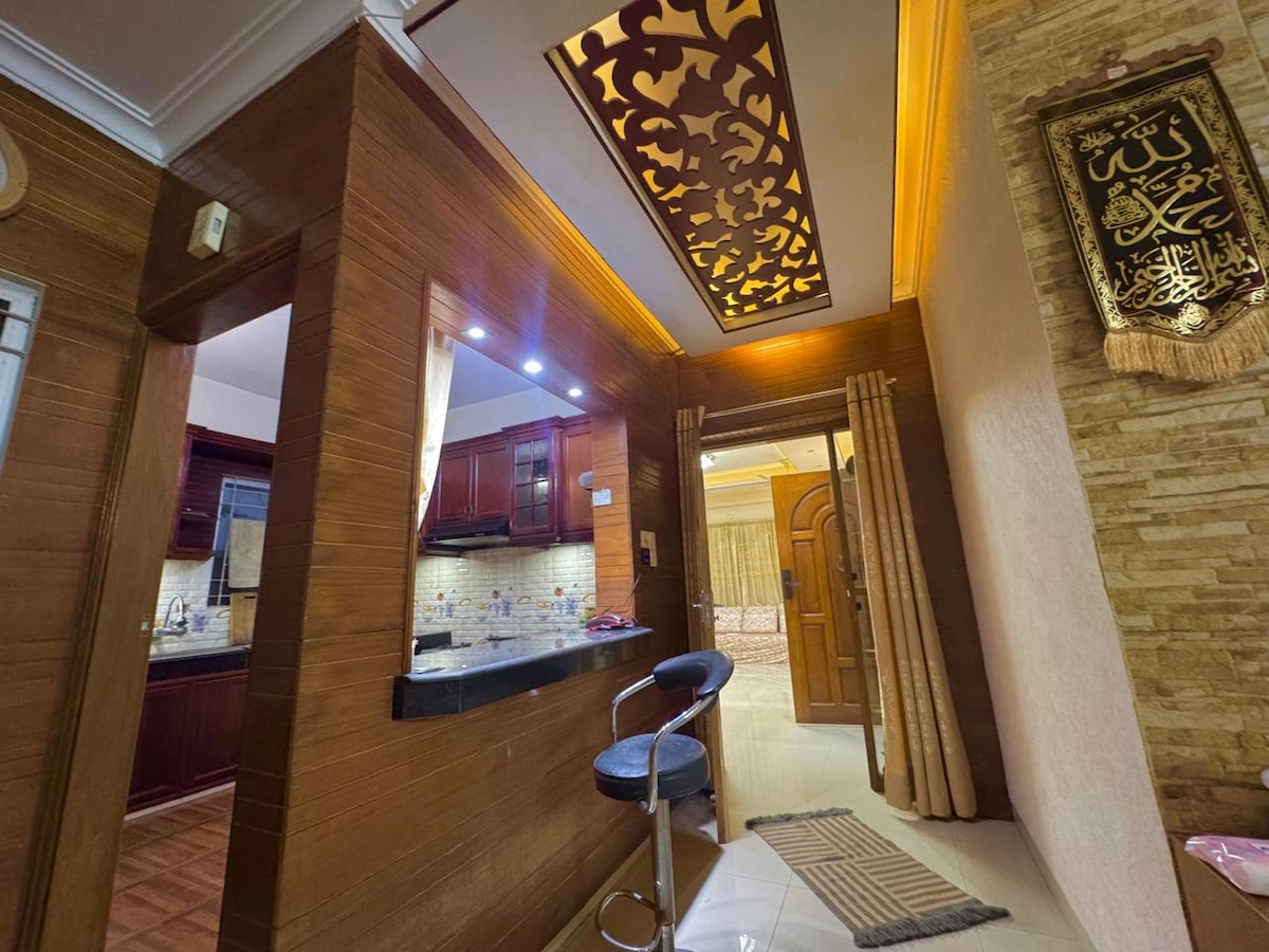 A modern kitchen area features dark wooden cabinetry and a granite countertop, illuminated by under-cabinet lighting. A bar stool is positioned at the counter, while decorative elements and a patterned ceiling draw attention to the room's design. Warm tones create a welcoming ambiance.