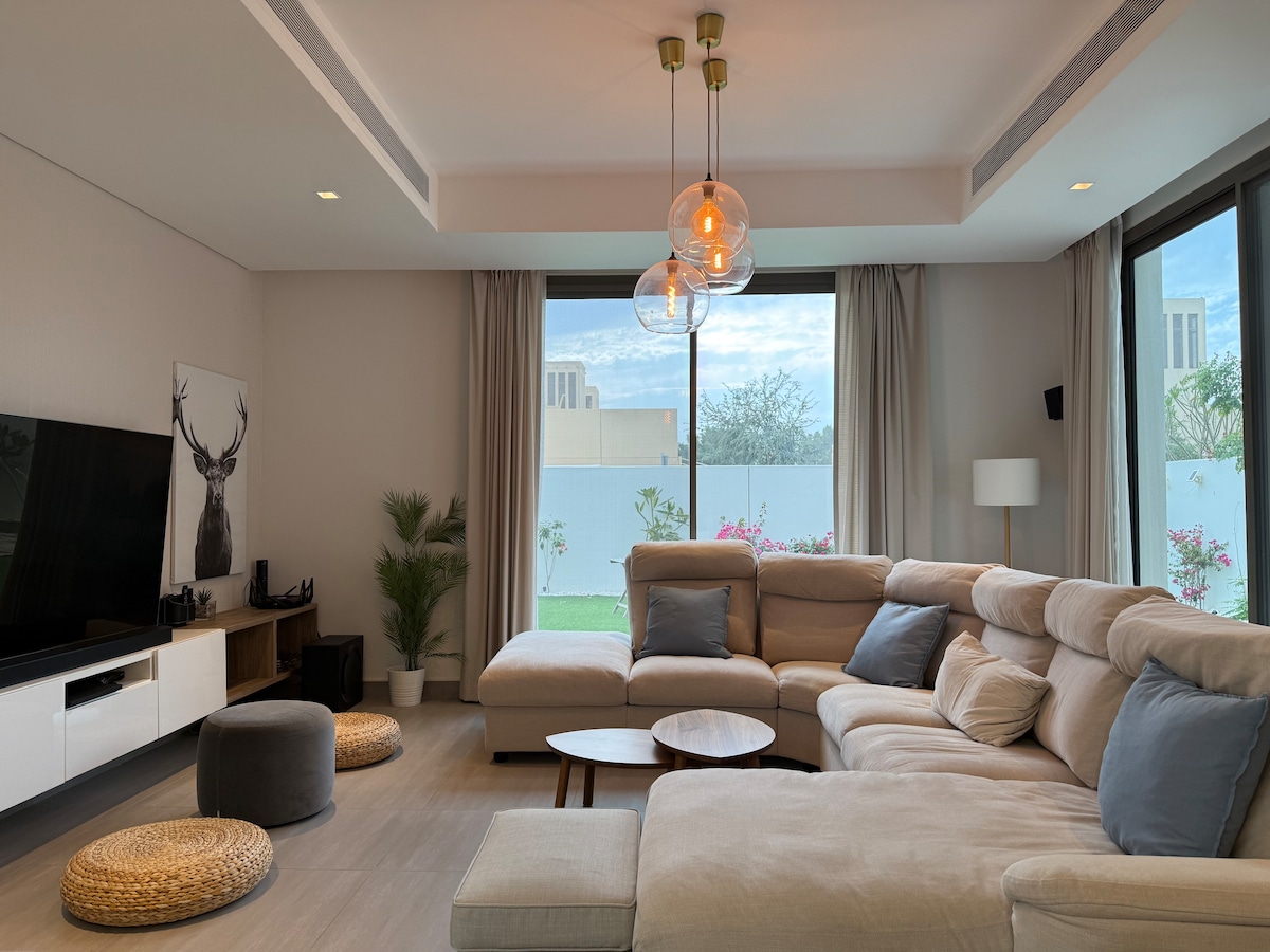 A spacious living area features large windows that allow natural light to illuminate the room. A sectional sofa, small round tables, and a variety of cushions create a comfortable seating arrangement. Decorative elements include a plant in the corner and modern light fixtures hanging from the ceiling.
