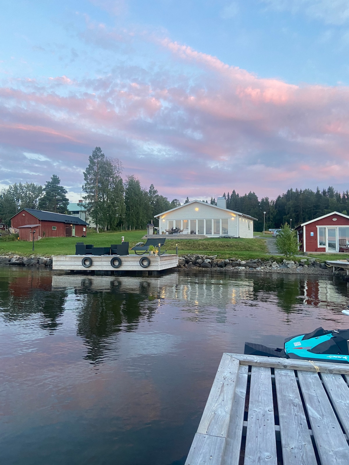 A serene lakeside scene is depicted, showing a modern white house next to the water, with large windows reflecting the sunset. Surrounding structures include traditional red cabins. The calm water mirrors the sky's soft colors, creating a peaceful ambiance.