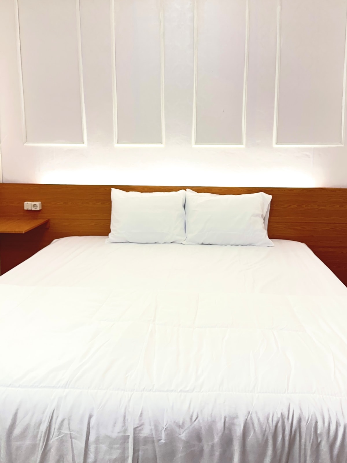 A spacious king-size bed is centered against a softly illuminated wooden panel wall. The bed features a fresh, white linen cover and two neatly arranged pillows. A wooden bedside table is positioned near the bed, contributing to the room's simple elegance.