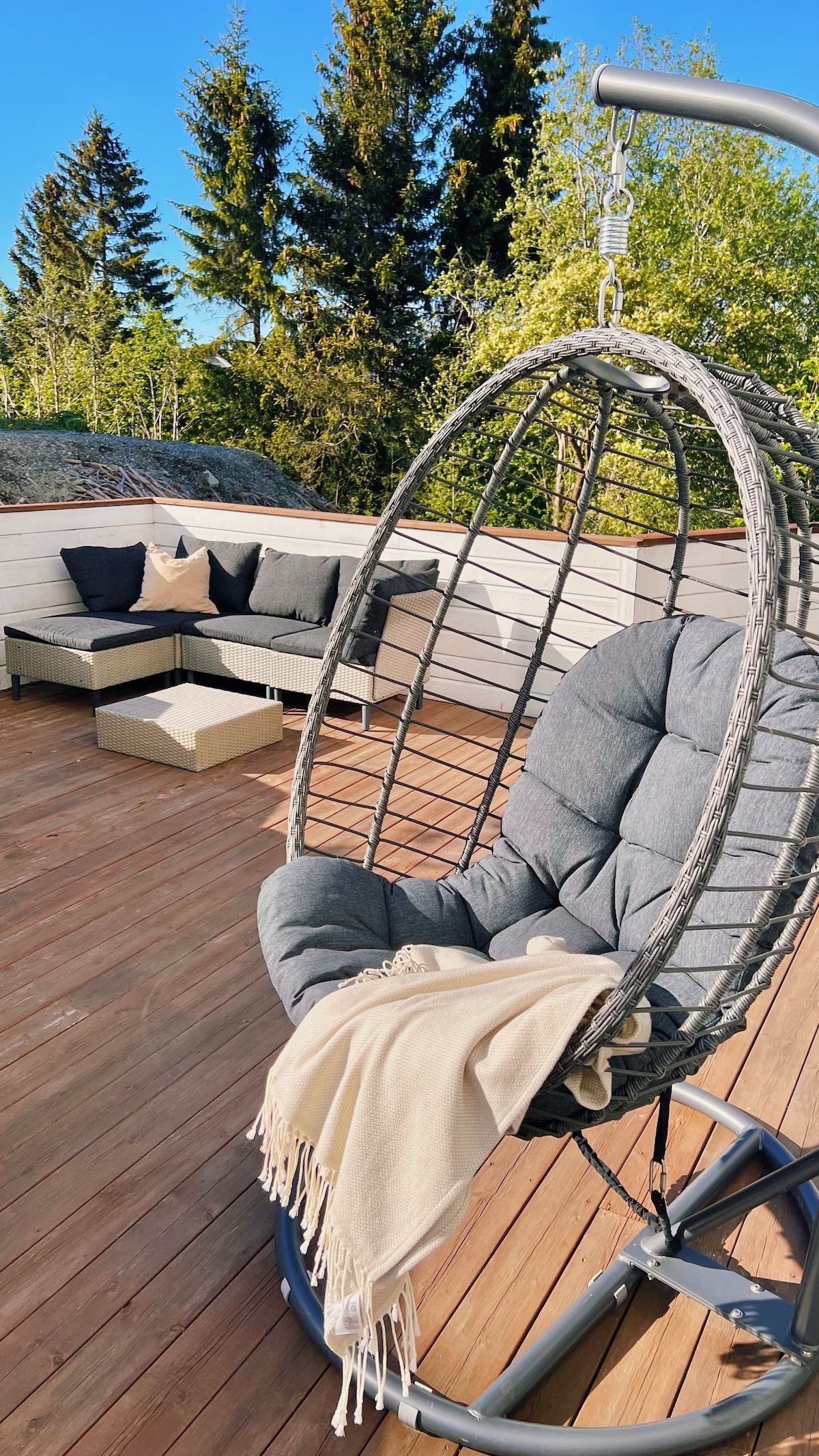A cozy terrace area features a suspended hanging chair with plush cushioning and a decorative throw draped over it. A modern seating arrangement with dark cushions is positioned nearby, surrounded by greenery. The wooden deck receives abundant sunlight, enhancing the outdoor space's inviting feel.