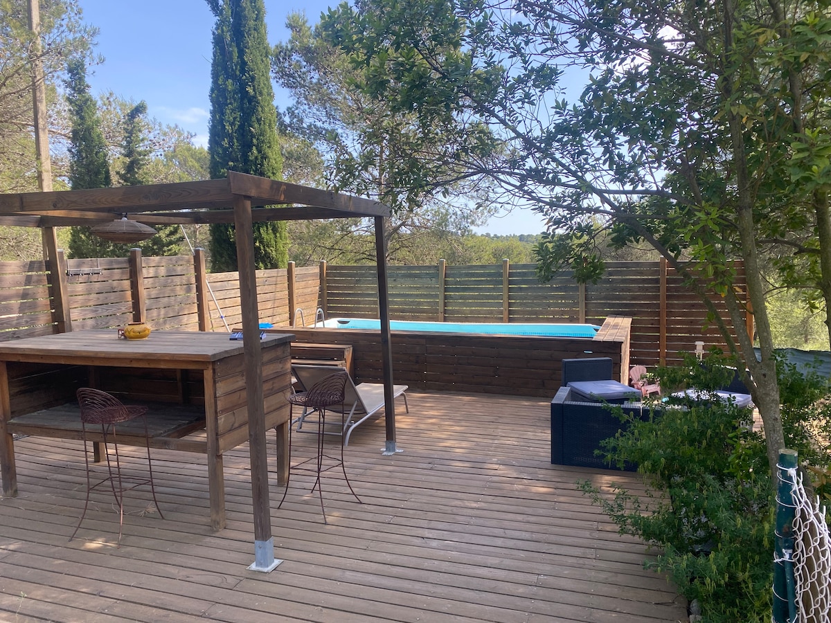 A wooden deck is visible, featuring a shaded outdoor dining area with two chairs and a table. A private pool can be seen in the background, surrounded by greenery and a wooden fence, offering a sense of privacy and tranquility.