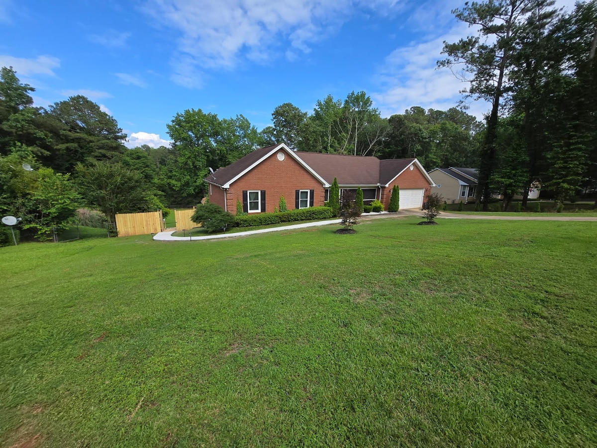 A brick home is set in a landscaped yard, bordered by greenery and trees. A circular driveway leads to the entrance, while a wooden fence outlines the property. The sky is clear with scattered clouds, creating a bright and welcoming atmosphere.