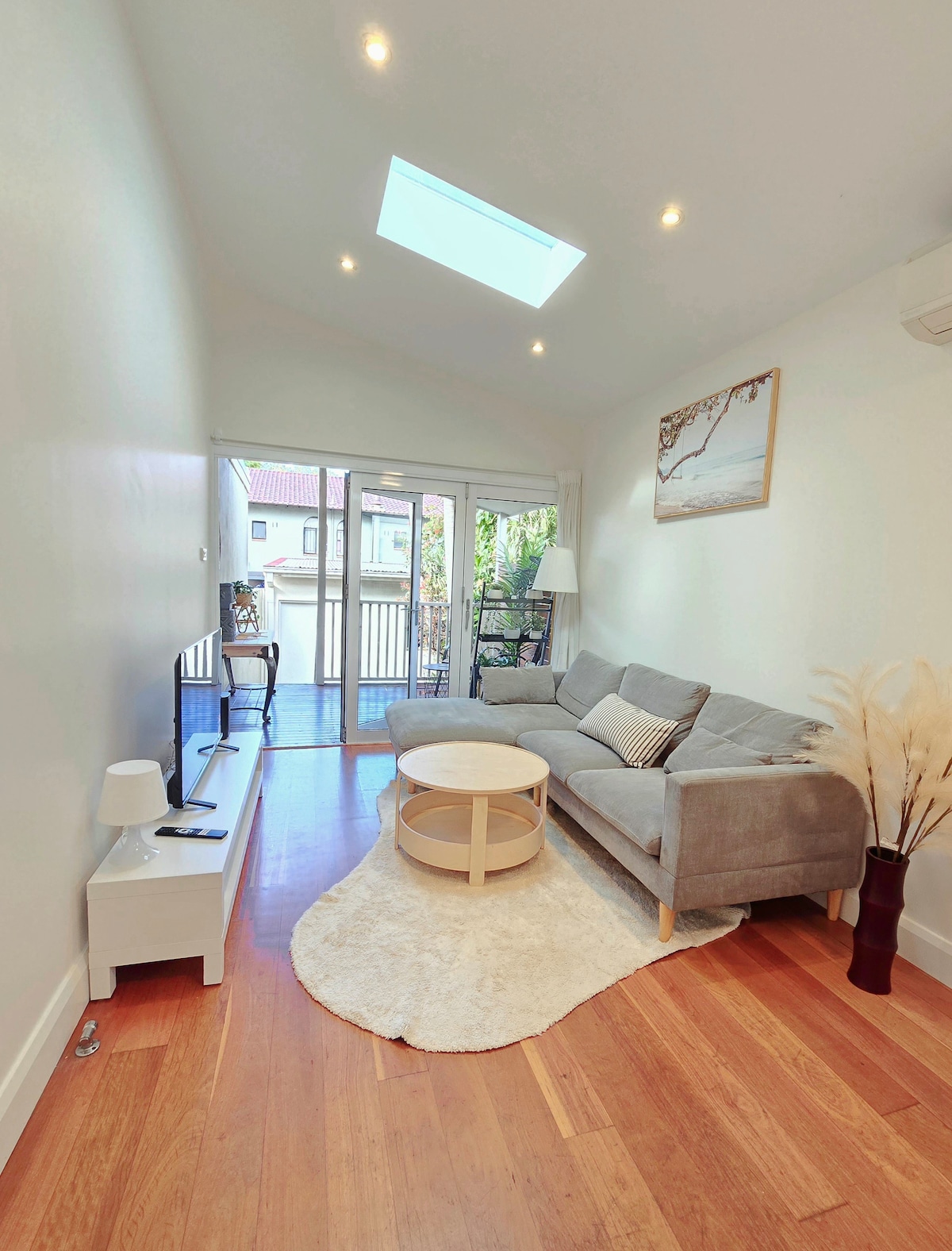A high-ceiling living room is observed, featuring a skylight that enables natural light to fill the space. A comfortable gray sofa is positioned beside a round coffee table on a soft area rug. Sliding doors lead to an outdoor deck area, enhancing the connection with nature.