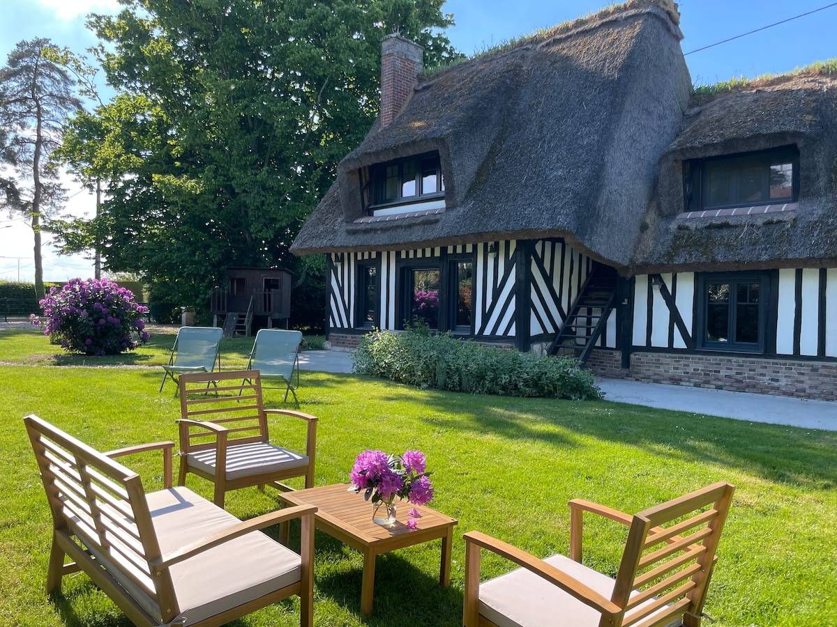 A charming thatched-roof cottage is set within a well-maintained garden. Comfortable outdoor seating arrangements are placed on the lush green lawn, complemented by a small table adorned with flowers. The exterior features traditional timber framing, and pleasant greenery surrounds the property.