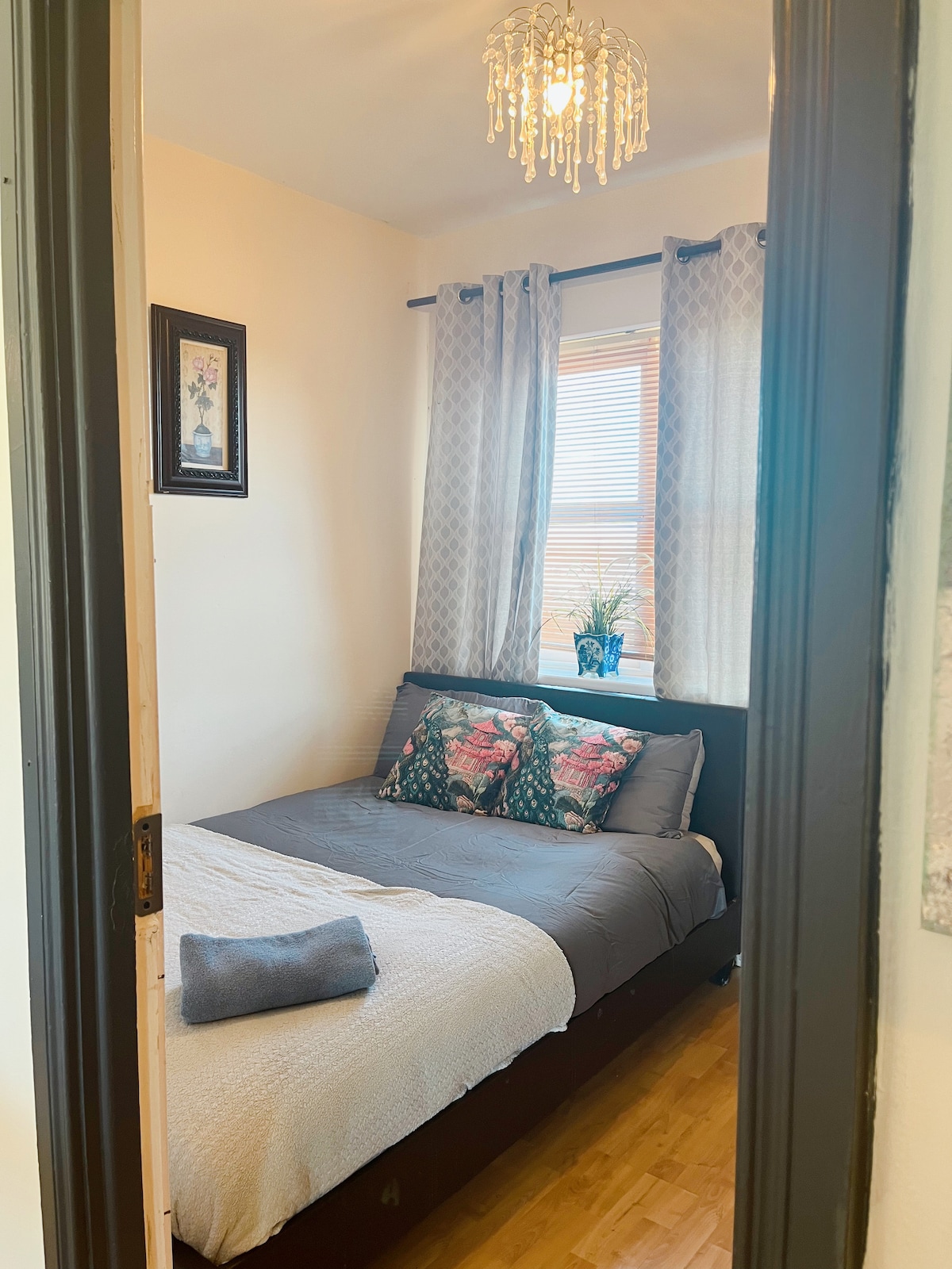 A bedroom is captured from the doorway, featuring a spacious bed adorned with decorative pillows. Soft curtains frame a window that allows natural light to fill the room. A small potted plant sits on the windowsill, and a crystal chandelier adds elegance to the ceiling.