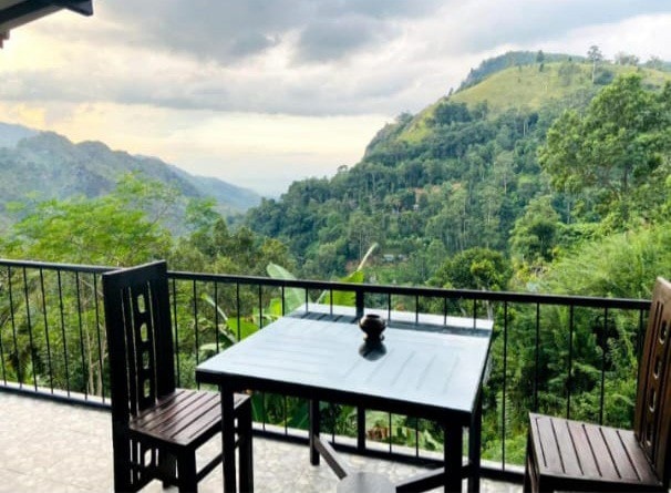 A balcony offers a table and two chairs, set against a backdrop of lush green hills and mountains. The landscape appears vibrant and expansive, with varying shades of greenery under a cloudy sky.