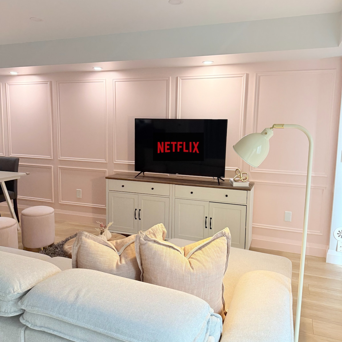 A modern living area features a light-colored sofa with decorative pillows, positioned towards a flat-screen television displaying the Netflix logo. The wall behind is finished with soft pink paneling, and a sleek floor lamp adds illumination to the space.