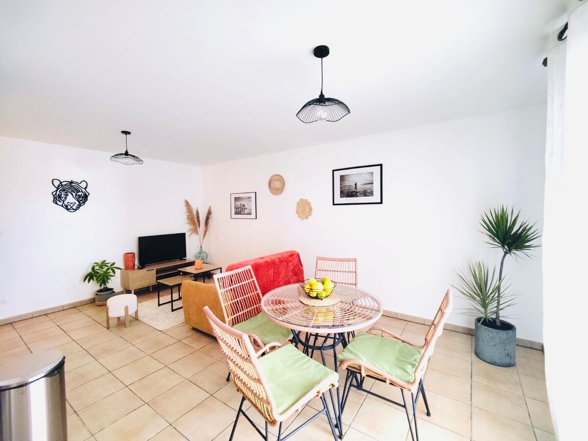 A bright living area is presented, showcasing a round dining table surrounded by four woven chairs. A comfortable sofa and a television are visible, with decorative plants and wall art enhancing the space. Natural light enters through the windows, contributing to a welcoming atmosphere.