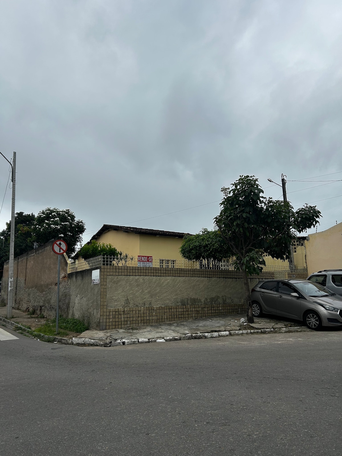 A simple yellow house is visible from the street, framed by a wrought iron fence. Lush green trees provide some shade, while parked cars are seen along the curb. The sky is overcast, creating a muted atmosphere around the property.