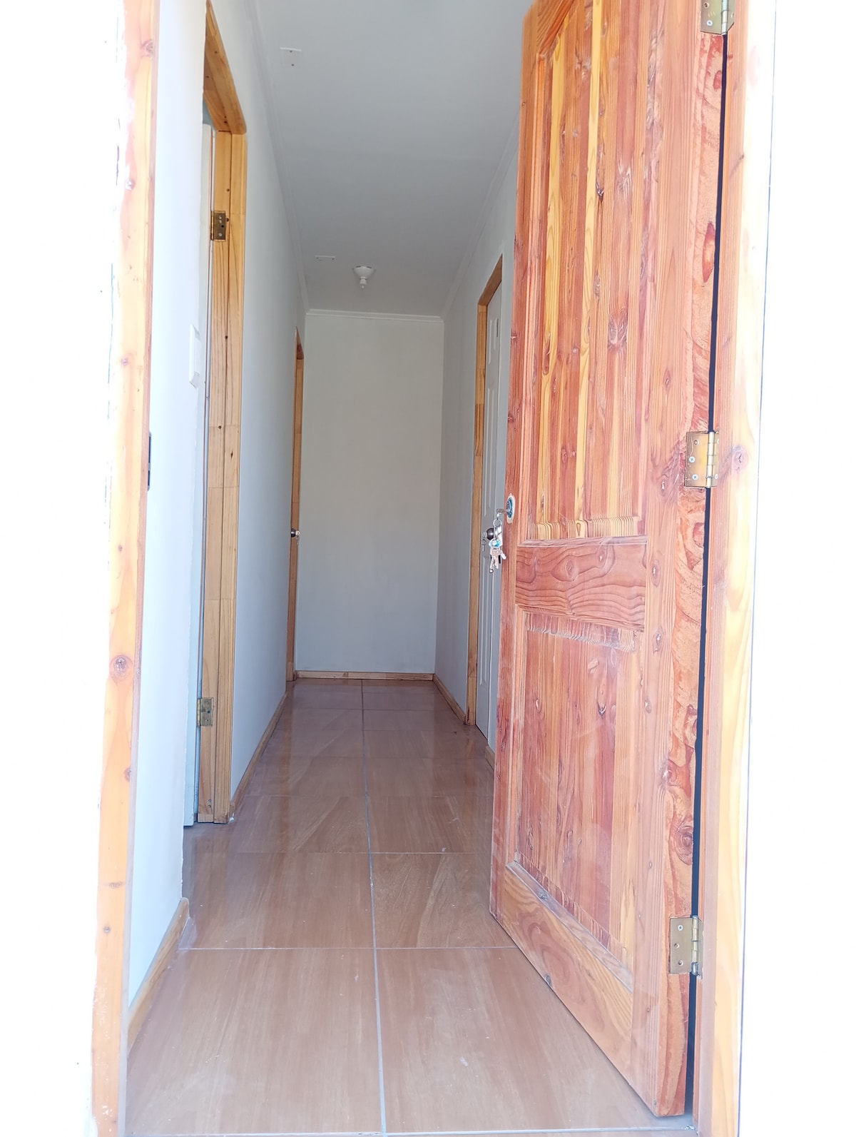 A hallway is depicted, featuring natural wood doors on either side. The floor is tiled and reflects natural light, while the walls are painted a neutral color, contributing to a sense of openness. The space appears well-maintained and accessible.