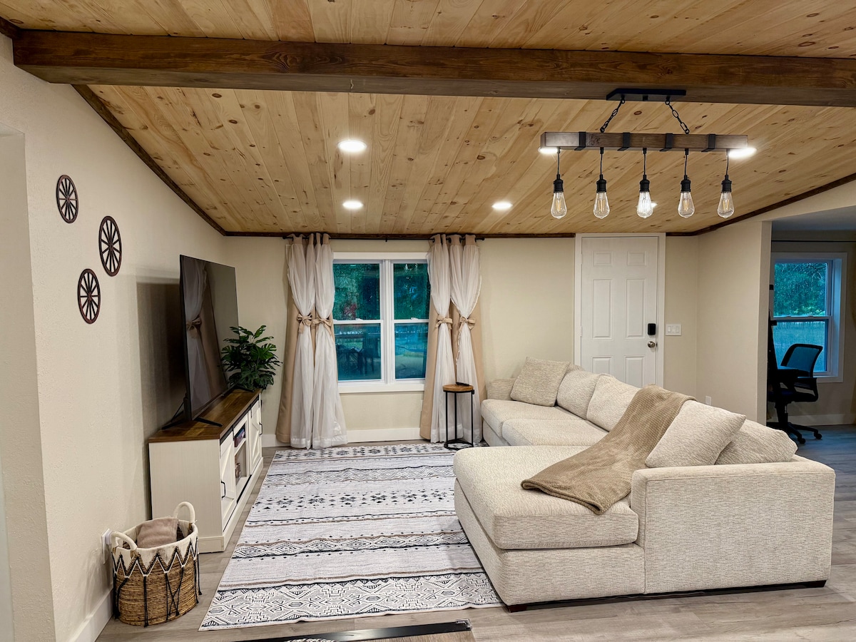 A spacious living area features a light-colored sectional sofa, accented with a soft throw blanket. Bright, airy curtains frame the windows, which overlook the outdoor space. A television is mounted on a modern cabinet, and decorative wall elements add a touch of character.