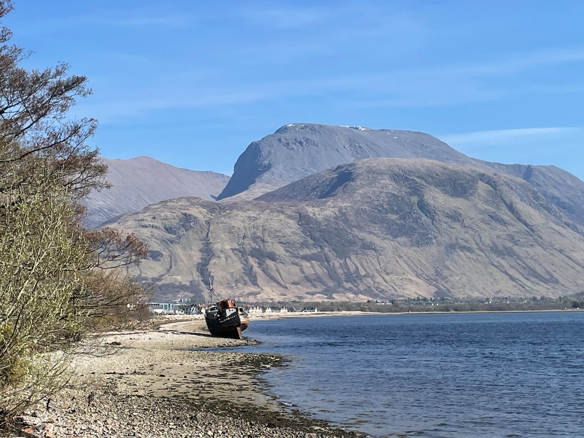 A scenic view captures a shipwreck resting on the shoreline, surrounded by a pebbled beach. In the background, the majestic Ben Nevis dominates the landscape, with its peaks partially covered in snow under a clear blue sky. The tranquil water reflects the surrounding natural beauty.
