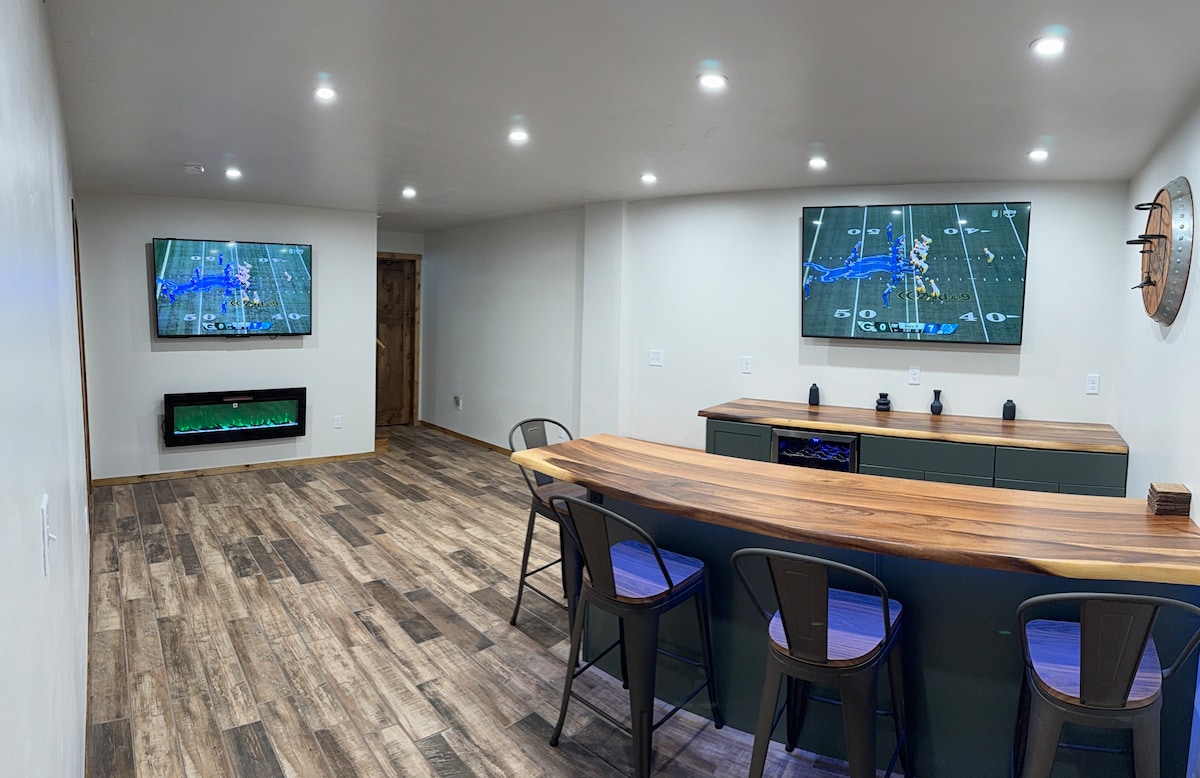 A spacious recreational area features warm wooden flooring and well-lit walls. Two large screens are mounted on opposite walls, displaying sports events. A stylish bar counter is positioned against the wall, accompanied by several metal stools, providing a comfortable spot for relaxation.
