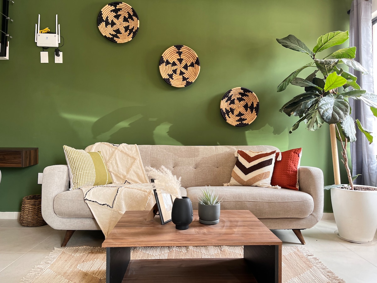 A comfortable living area features a light gray sofa adorned with accent cushions in warm tones. A wooden coffee table sits in front, displaying a small plant and a decorative object. Woven baskets are arranged on the green wall, adding texture and interest.