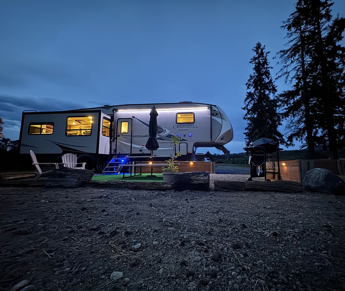 An RV is positioned on a gravel and grass lot, illuminated softly under the evening sky. Steps lead up to the entrance, while outdoor seating and a grill are present. Tall trees frame the background, completing the serene nature setting.