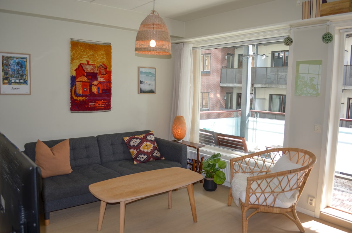 A cozy living area features a grey sofa adorned with colorful cushions. A light wood coffee table sits in front. Natural light floods the room through large windows, showcasing a view of the balcony. A rattan armchair adds a touch of warmth, complemented by wall art.
