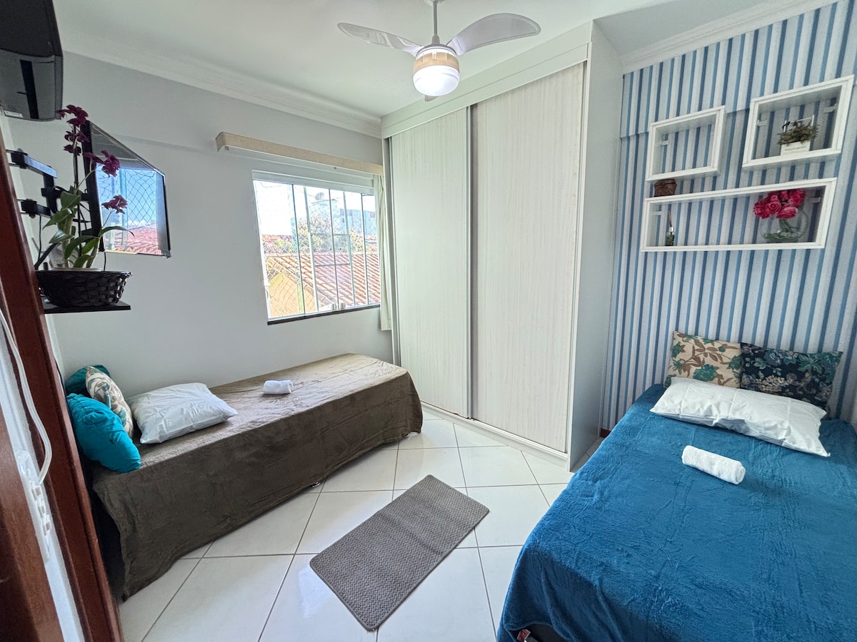 A well-lit bedroom features two single beds, one with a blue bedspread and a white pillow. A tan bed is adjacent to it, accompanied by a cushioned mat. A window allows natural light in, and a white closet occupies one wall, with decorative shelves holding potted plants.