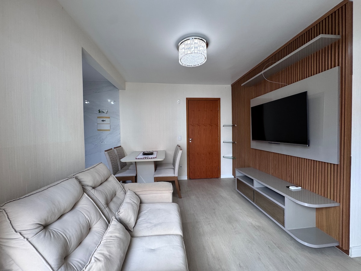 A cozy living area features a comfortable sofa and a sleek entertainment unit with a flat-screen TV mounted on the wall. A dining table is positioned nearby, providing seating for four. Natural light fills the space through a window, enhancing the light-colored walls.