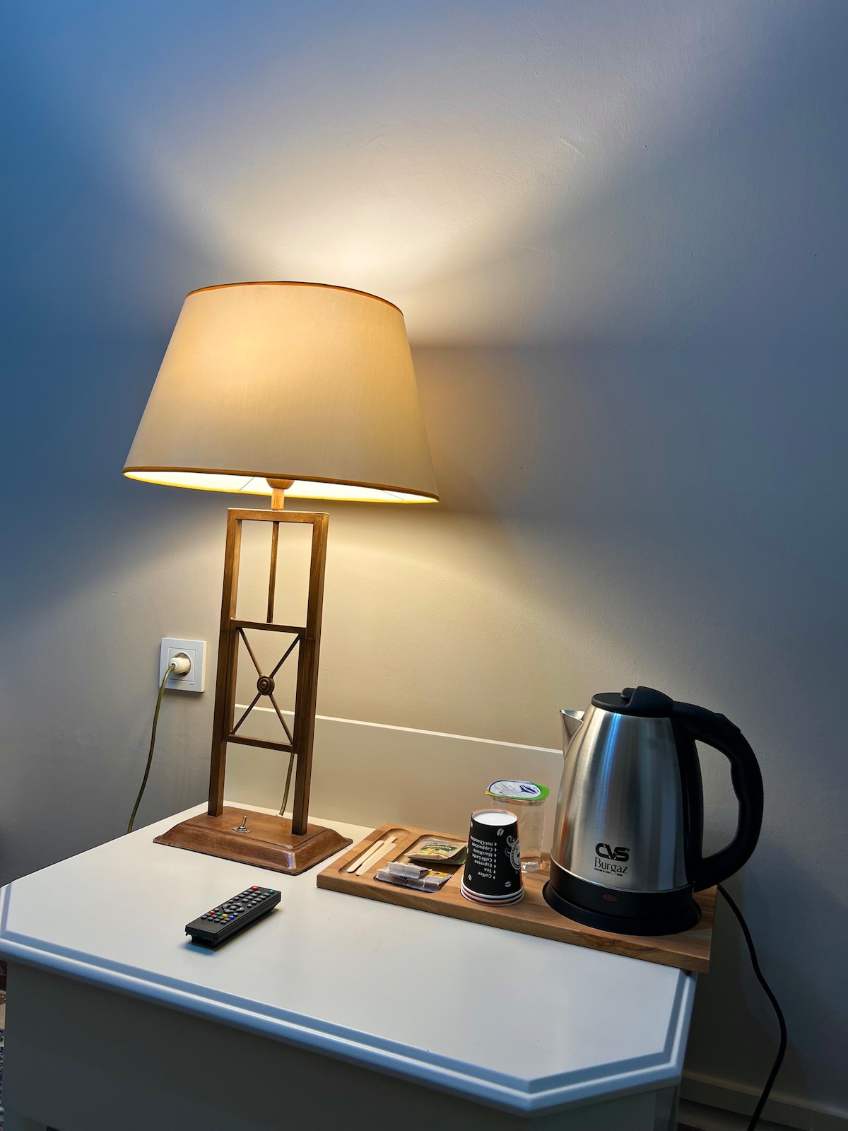 A bedside table is equipped with a modern lamp casting a warm glow. A stainless steel electric kettle sits alongside complimentary tea and coffee supplies on a wooden tray. A remote control lies on the table, providing convenience for guests.