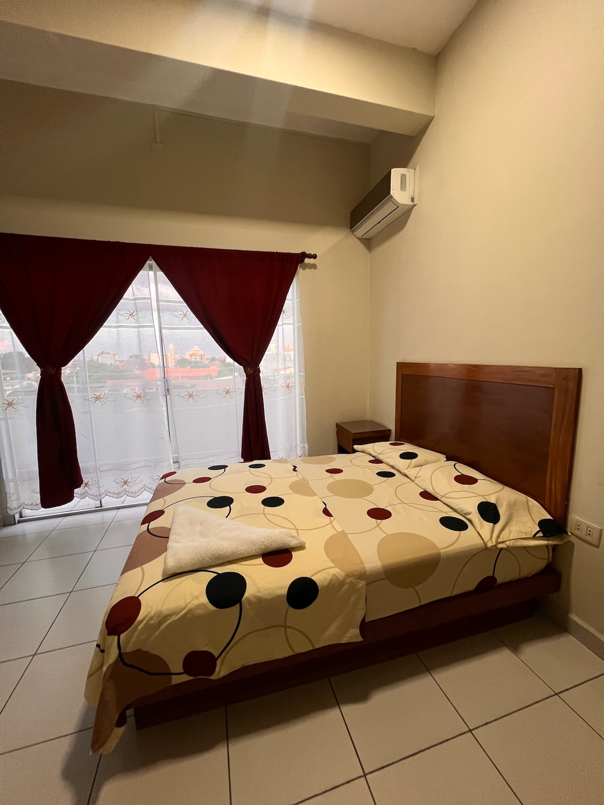 A bright bedroom is equipped with a spacious bed adorned with a patterned blanket featuring polka dots. Light enters through a large window dressed in sheer curtains, complemented by heavy drapes in a rich color. A bedside table is positioned next to the bed.