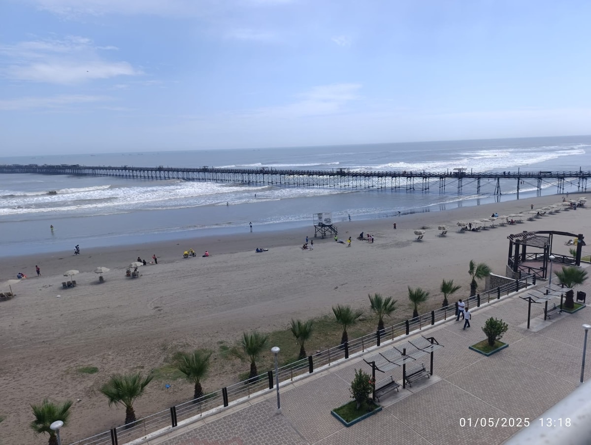 A scenic view of a sandy beach is presented, featuring gentle waves lapping at the shore. In the distance, a long wooden pier extends over the water. Sun loungers and palm trees are placed along the beachfront, creating a relaxed coastal atmosphere.