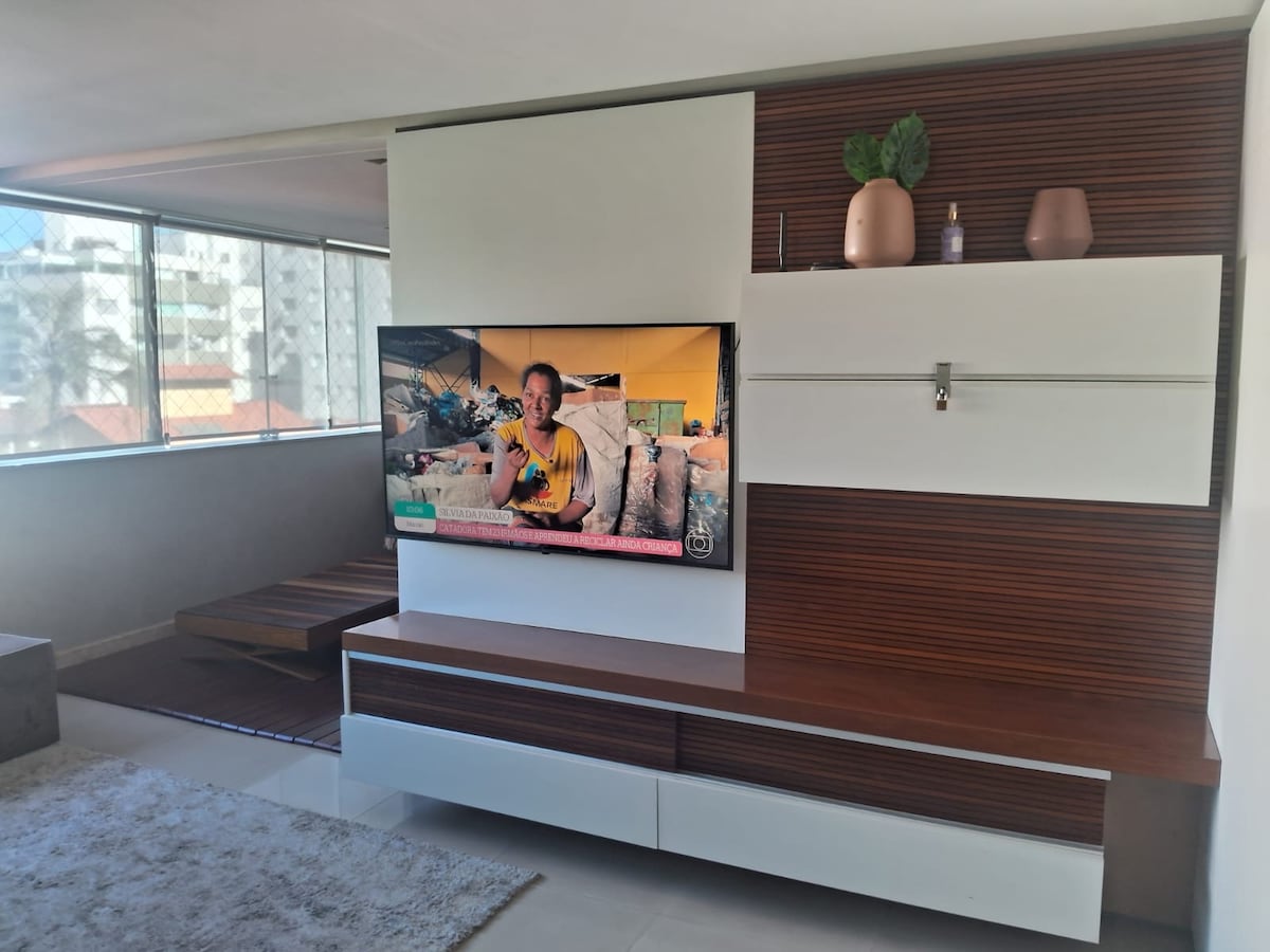 A modern living area is displayed, featuring a mounted television on a sleek wall unit with a combination of white and wood finishes. Natural light enters through large windows, highlighting a neutral-toned rug and a simple coffee table. Decorative plants and items are placed on the unit.