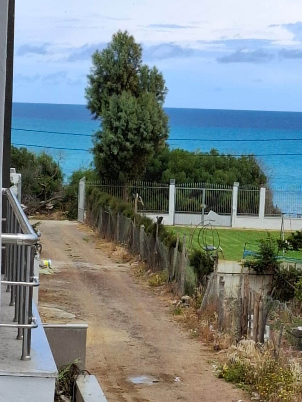 A dirt pathway leads towards a grassy area bordered by a white fence. Beyond the fence, a vibrant blue ocean is visible under a cloudy sky. A tall tree is positioned to the left, adding greenery to the coastal scene.