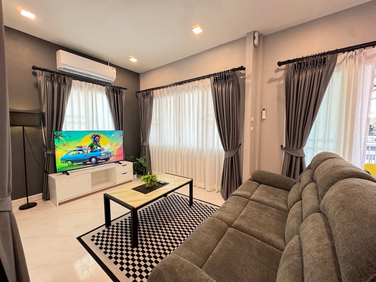 A modern living room features a comfortable gray sofa positioned opposite a large television set. Natural light filters through two windows adorned with sheer white curtains. A small green plant is placed on a rectangular coffee table, set on a patterned black and white rug.