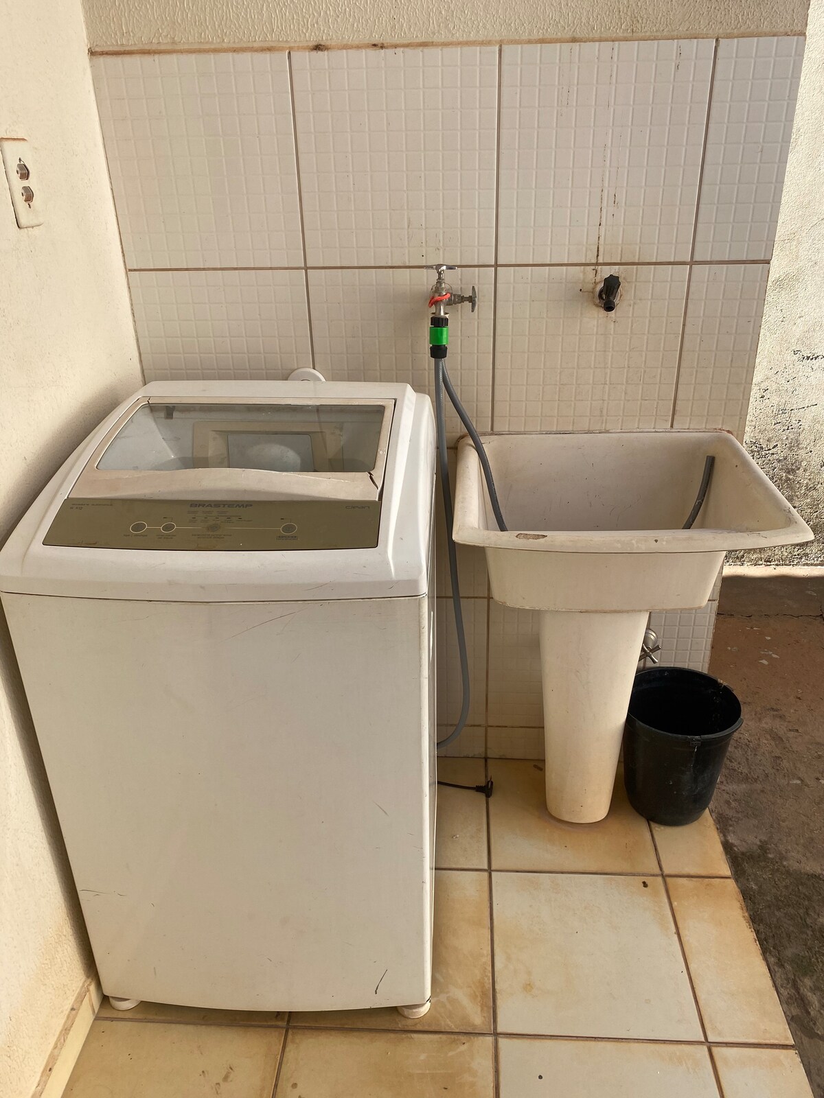 A compact laundry area is presented featuring a washing machine alongside a utility sink. The walls are tiled in light colors, and a hose connects the sink to the water supply. A small black bucket is positioned on the floor nearby.