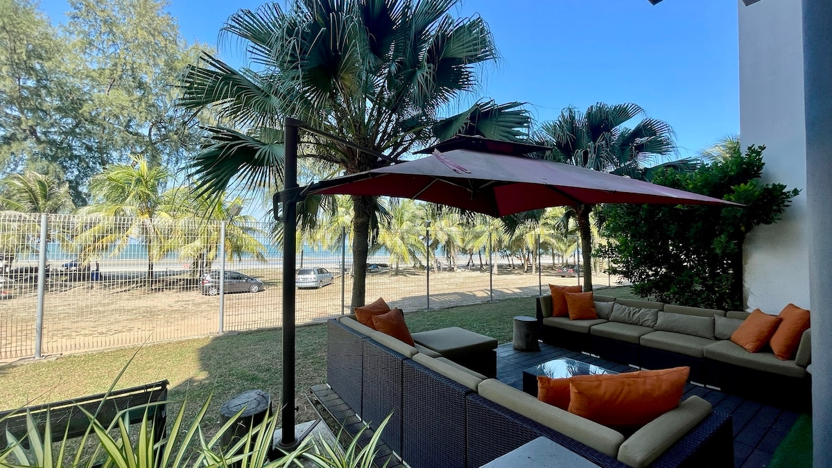 An outdoor seating area features a large, shaded sofa set with orange accent pillows. A coffee table sits in the center, surrounded by lush greenery and palm trees. The ocean can be seen in the background, providing a relaxed coastal atmosphere.