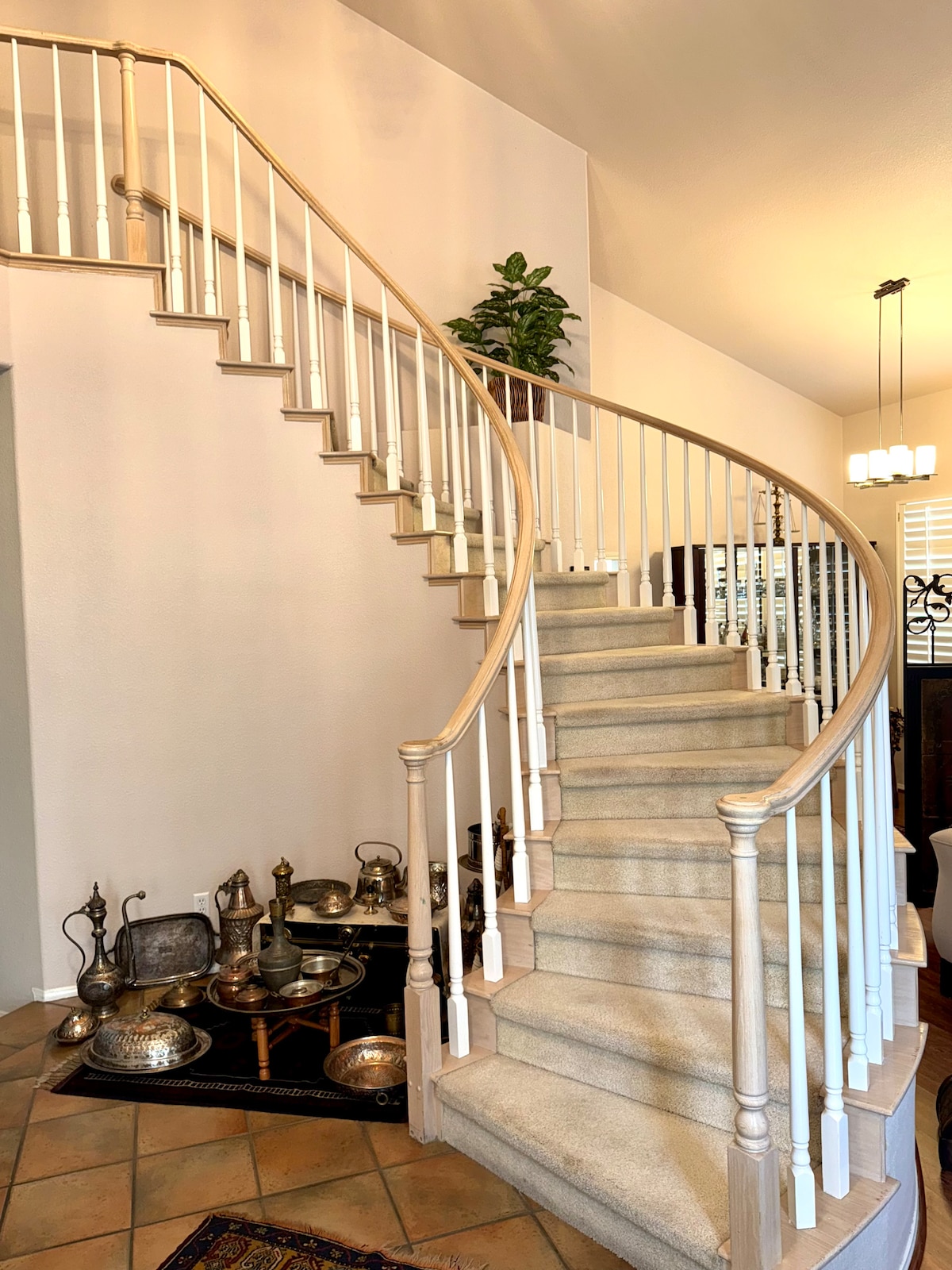 A gracefully curved staircase is seen, featuring a smooth handrail and light-colored carpeting. At the bottom, a collection of metallic artifacts is arranged on the floor, adding a touch of character to the space. Natural light pours in from the ceiling fixtures above.