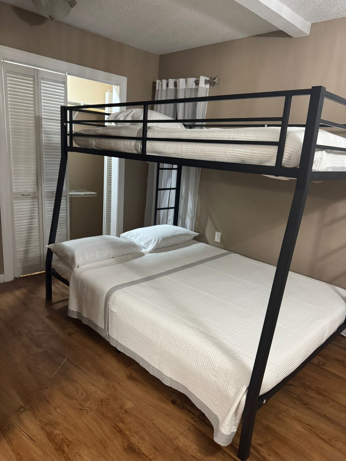 A furnished sleeping area features a sturdy metal bunk bed above a spacious lower bed, both dressed in light-colored linens. Natural light filters through an adjacent window, while a closet with sliding doors is visible in the background.