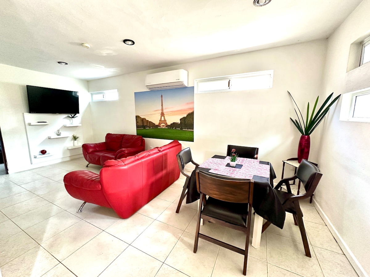 A modern living area is presented with a red sectional sofa and a dining table set for four. A wall-mounted television is positioned opposite the seating area. Sunlight enters through several windows, and a large indoor plant adds a touch of greenery to the space.