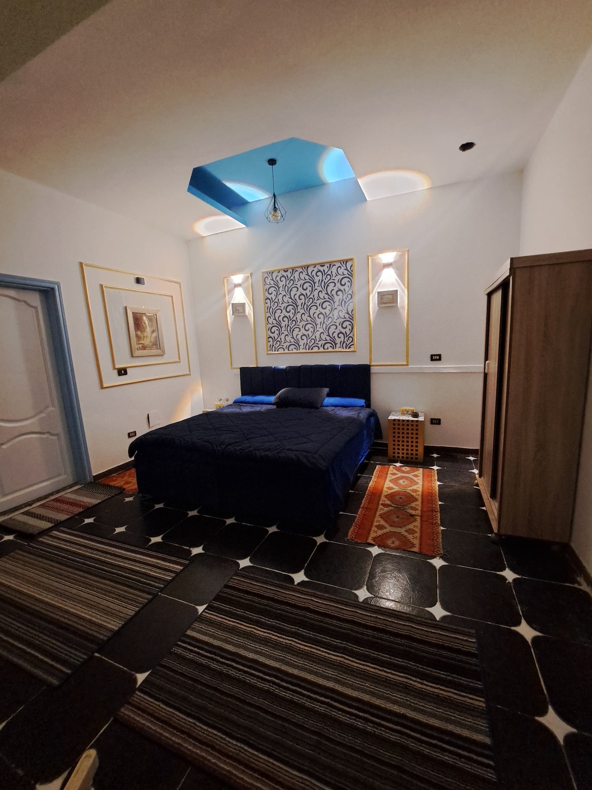 A spacious bedroom features a large, neatly made bed with blue bedding. Decorative wall artwork is positioned above the headboard, and stylish light fixtures are mounted on either side. A patterned rug covers the floor, while a wardrobe is visible in the corner.