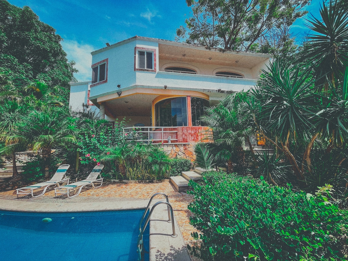 The residence features a multi-level structure surrounded by lush greenery. A private pool is visible in the foreground, bordered by lounge chairs. A spacious patio connects the house to the garden, with large windows allowing natural light to fill the interior.