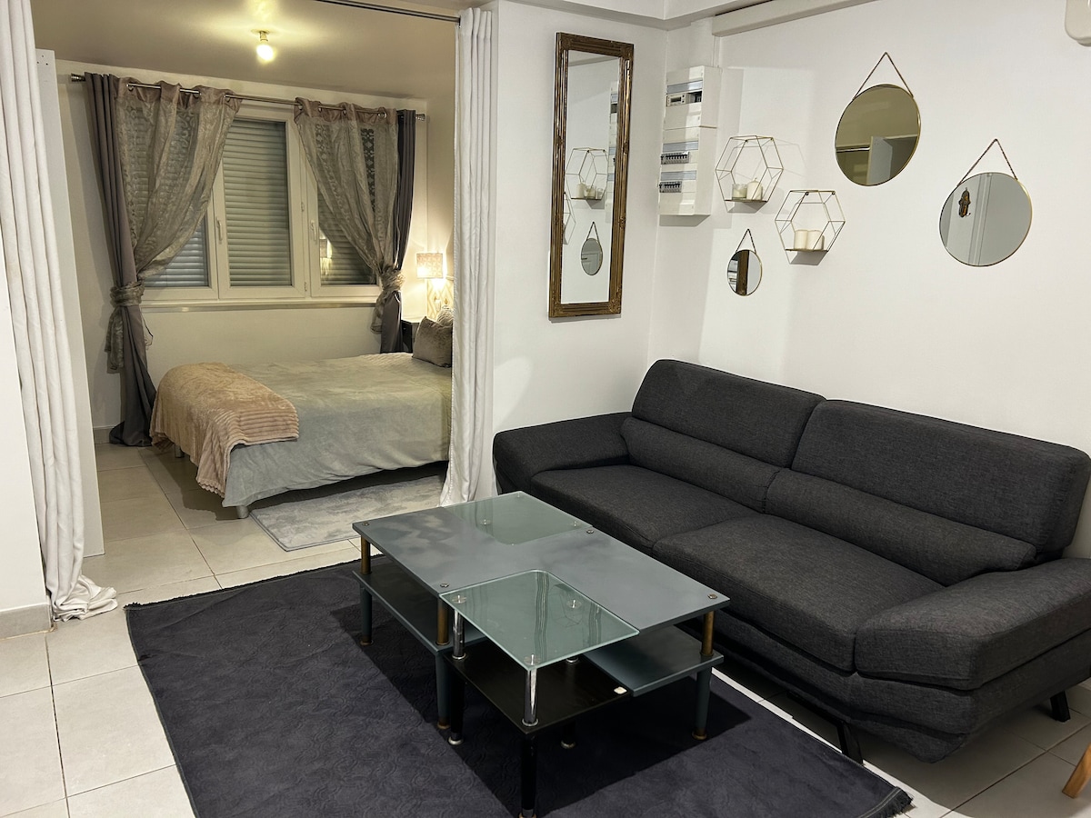 The open living space features a dark gray sectional sofa adjacent to a glass coffee table. A soft area rug lies beneath, while a cozy bedroom with a large bed and decorative wall mirrors can be seen through the space, offering a well-lit ambiance.