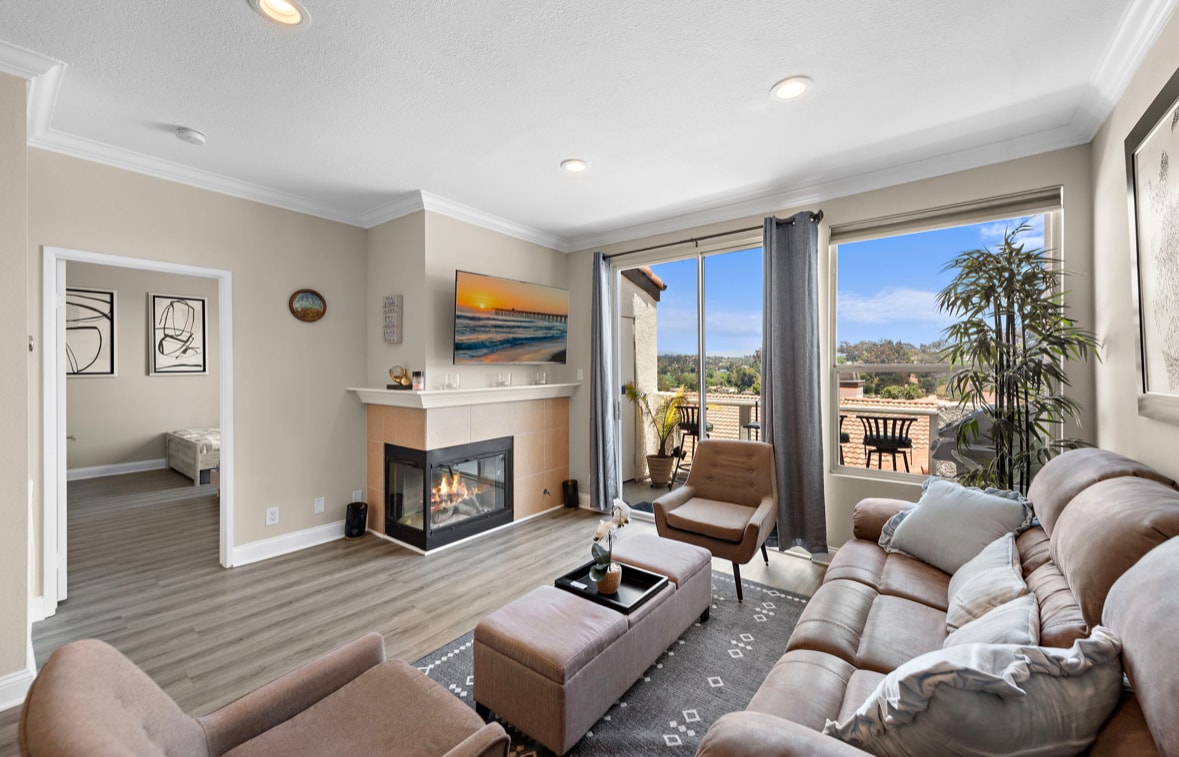 A cozy living area features a fireplace and large windows that provide ample natural light and views of the outside. Comfortable seating is arranged around a central coffee table, with subtle decor enhancing the inviting ambiance. A balcony is visible, offering an outdoor seating area.