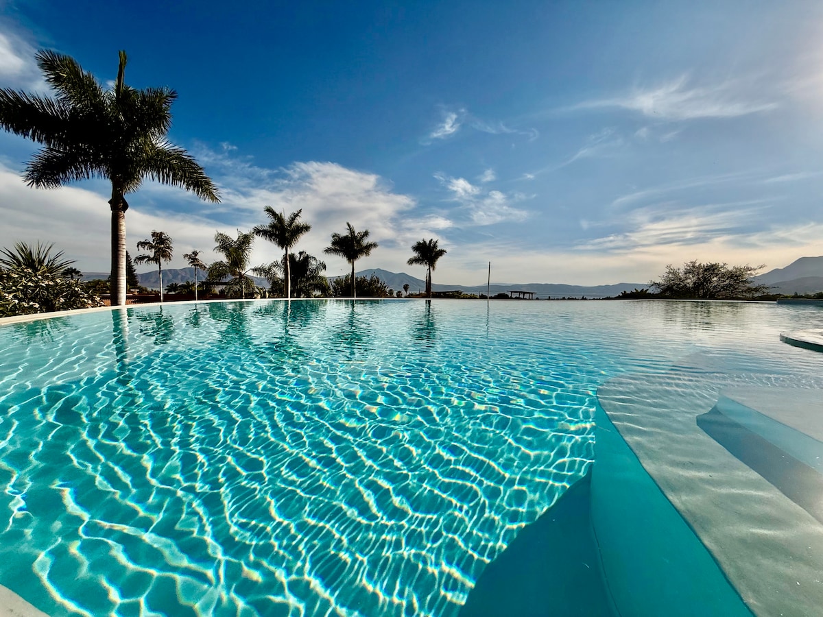 A serene infinity pool reflects the sky, showcasing shimmering water under bright sunlight. Palm trees line the poolside, enhancing the calm atmosphere. Subtle mountain views are visible in the background, contributing to the peaceful setting.