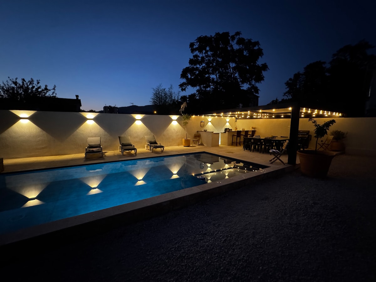 The night view presents a serene pool area, with the water reflecting warm light from surrounding fixtures. Comfortable lounge chairs are placed beside the pool, while a shaded dining space is illuminated by string lights, creating a tranquil ambiance for outdoor relaxation.
