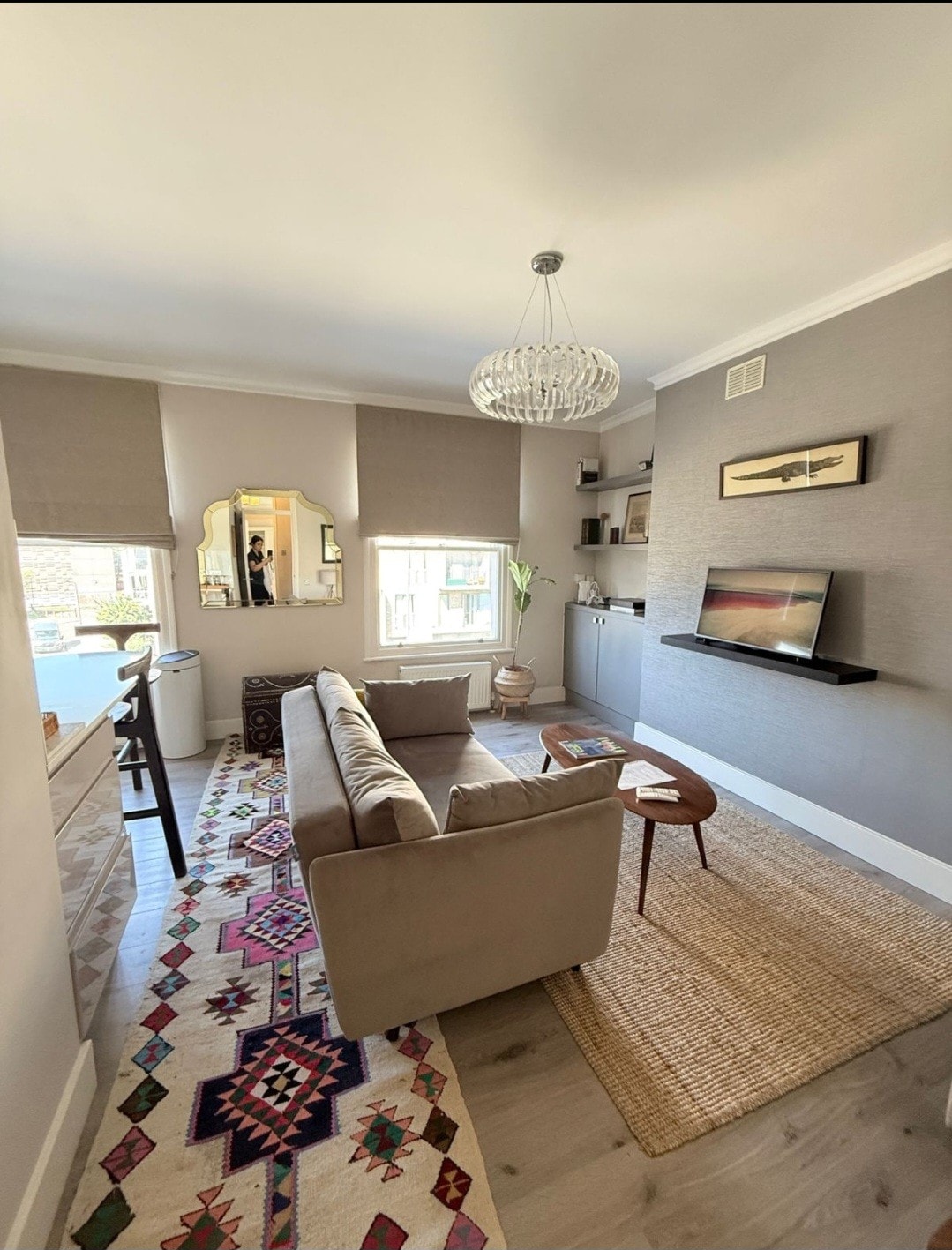 A living area features a comfortable sofa facing a wall-mounted TV. Light streams in through large windows adorned with shades. Decorative elements include a mirror, artwork on the walls, and a patterned rug underfoot, contributing to a cozy and inviting atmosphere.