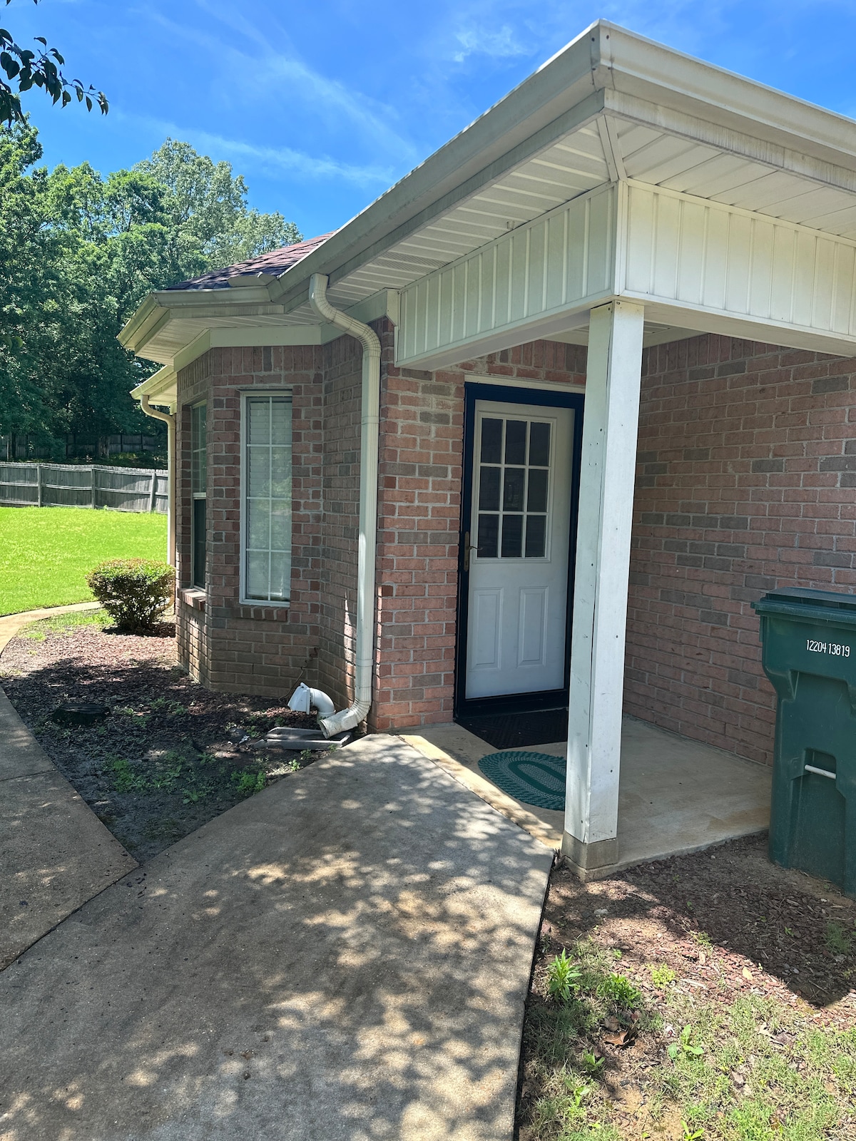 The exterior features a brick facade with large windows providing natural light. A covered entryway leads to a white front door, and a small porch area is visible. Green grass surrounds the home, framed by a walkway and a trash bin nearby.